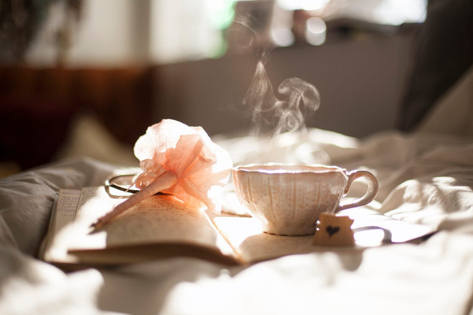 A steaming teacup beside soft pink flowers and an open notebook